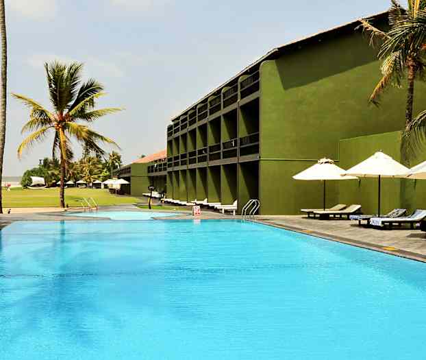 Ulkouima-allas hotellin vieressä, jossa on vihreitä rakennuksia, ympärillä palmuja ja aurinkotuoleja varjojen alla. (Tekoälyn luoma teksti)