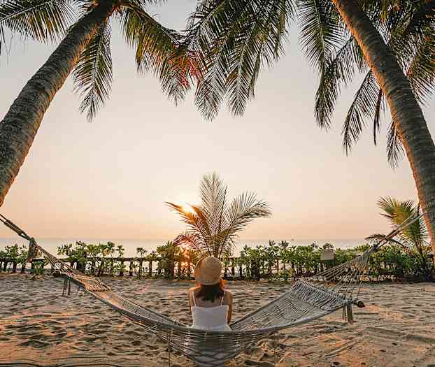 Henkilö istuu riippumatossa kahden palmun välissä rannalla, kun aurinko laskee horisontissa. (Tekoälyn luoma teksti)