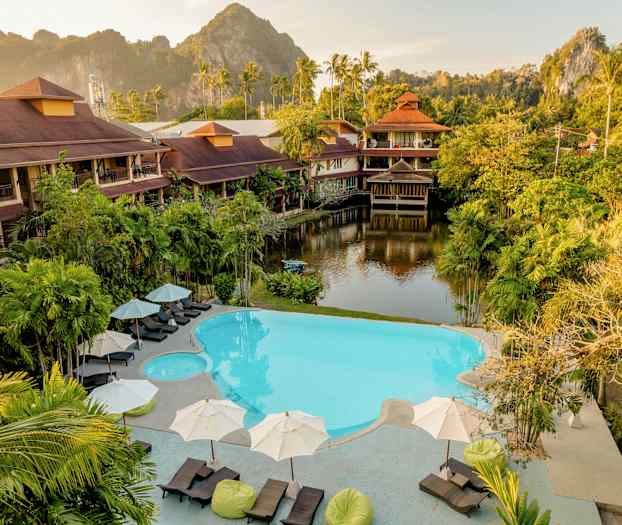 Hotellialue, jossa uima-allasalue, trooppisia kasveja ja perinteiseen tyyliin rakennettuja rakennuksia. Taustalla näkyy vehreä vuoristosiluetti. (Tekoälyn luoma teksti)