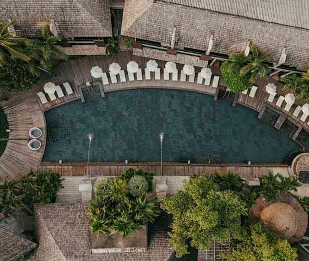 Ilmakuva uima-altaasta, jota ympäröivät aurinkotuolit, varjot ja vehreä kasvillisuus. (Tekoälyn luoma teksti)