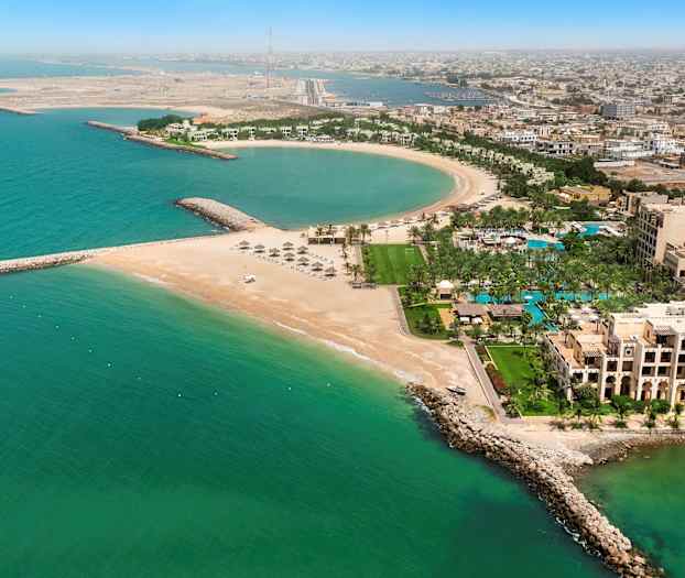 Ilmakuva rannikkokaupungista, jossa on puolikaaren muotoinen ranta, rakennuksia, palmuja ja turkoosia meren vettä. (Tekoälyn luoma teksti)