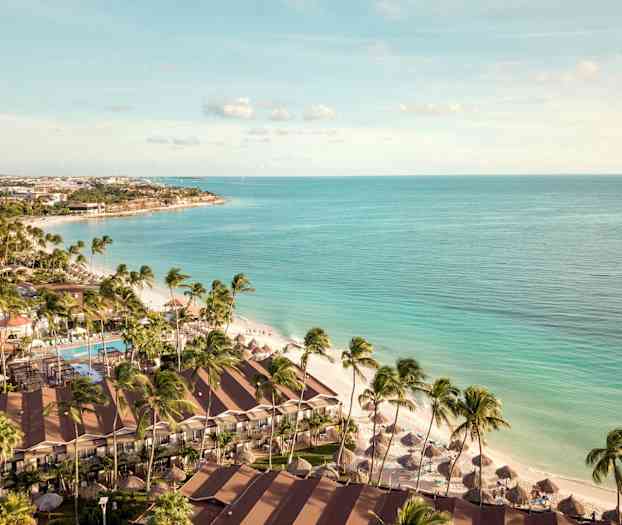 Ilmakuva rannasta, jossa on palmuja, hotellirakennuksia ja aurinkovarjoja kirkkaan sinisen meren lähellä. (Tekoälyn luoma teksti)