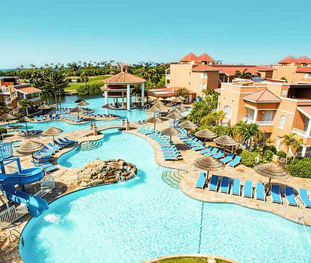 Kuva lomahotellista, jossa on allasalue, aurinkotuoleja ja aurinkovarjoja. Rakennukset ympäröivät aluetta, ja taustalla näkyy vihreitä palmuja. (Tekoälyn luoma teksti)