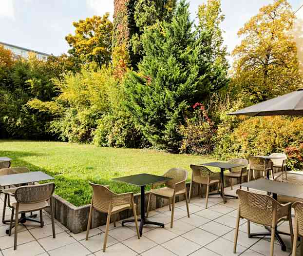 Ulkoravintola, jossa on pöytiä ja tuoleja laatoitetulla alueella viheralueen vieressä, jossa on pensaita ja puita. (Tekoälyn luoma teksti)