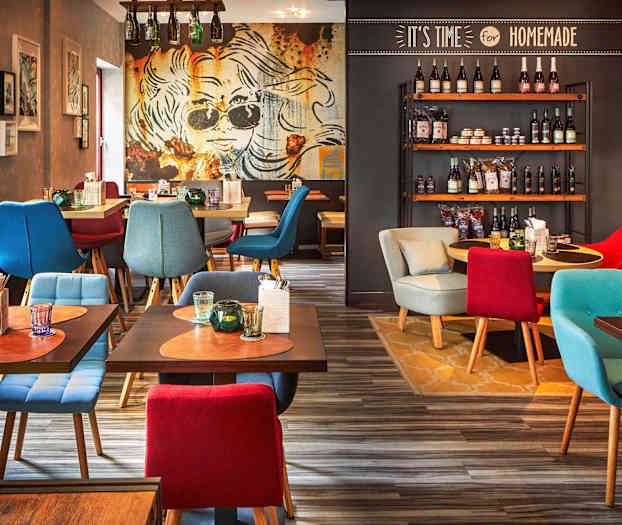 Ravintolasisustus, jossa värikkäitä istuimia, koristeellisia pöytiä juomilla ja aterimilla sekä seinä täynnä erilaisia tuotteita hyllyillä. (Tekoälyn luoma teksti)