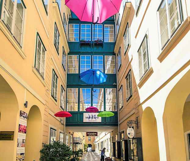 Kapea piha, jonka yläpuolella roikkuu värikkäitä sateenvarjoja, ja rakennuksien välinen kulkuväylä. (Tekoälyn luoma teksti)