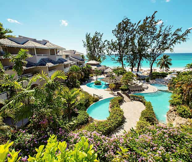 Kuva, jossa hotellirakennuksia ympäröi vehreys lähellä uima-allasta ja rantaa. (Tekoälyn luoma teksti)