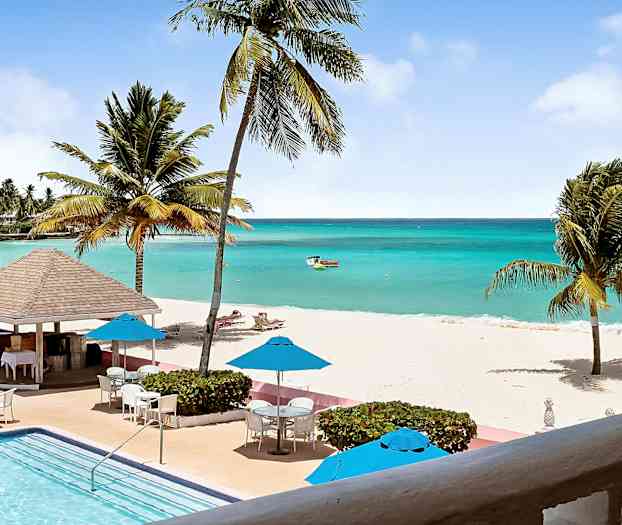 Trooppinen rantamaisema turkoosilla merellä, valkoisella hiekalla, palmuilla ja etualalla uima-altaalla. Aurinkovarjot ja ulkokalusteet sijaitsevat altaan vieressä. (Tekoälyn luoma teksti)