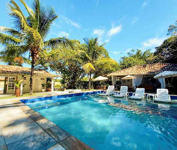 Uima-allas, jonka ympärillä on aurinkotuoleja, aurinkovarjoja ja palmuja rakennuksen vieressä aurinkoisessa säässä. (Tekoälyn luoma teksti)