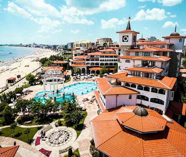 Lomakeskus, jossa on uima-allas, ympärillä punakattoisia rakennuksia, sijaitsee rannan vieressä, jolla on aurinkovarjoja ja aurinkotuoleja. (Tekoälyn luoma teksti)