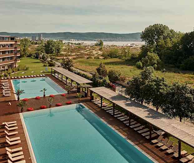 Suuri uima-allas, jonka vieressä aurinkotuoleja ja varjoja hotellin vieressä, taustalla vehreää maisemaa. (Tekoälyn luoma teksti)