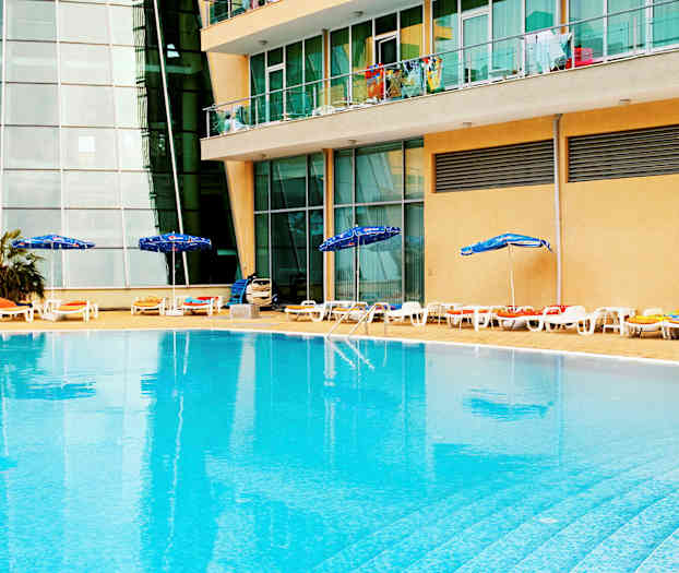 Ulkouima-allas, jonka reunalla on aurinkotuoleja ja aurinkovarjoja, sijaitsee rakennuksen edessä, jossa on lasi-ikkunoita ja parvekkeita. (Tekoälyn luoma teksti)