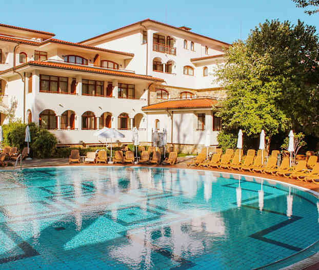 Uima-allas valkoisen rakennuksen edessä, jossa on punaiset katot, ympäröitynä aurinkotuoleilla ja vihreillä puilla. (Tekoälyn luoma teksti)