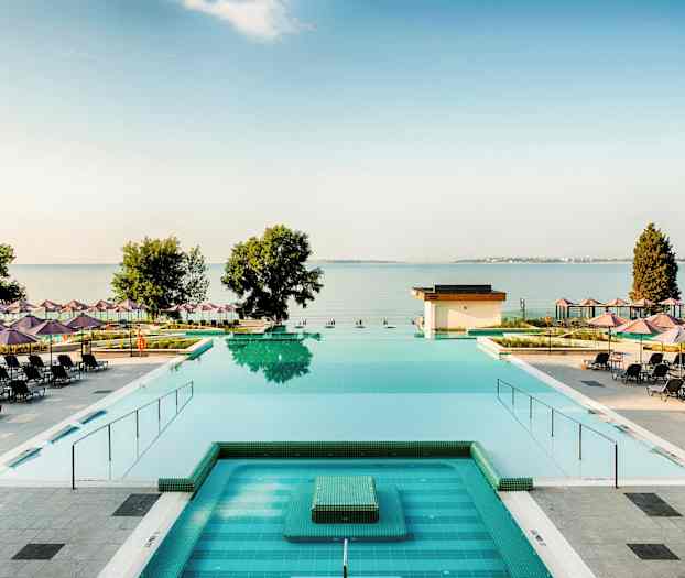 Näkymä uima-altaalle, jota ympäröivät aurinkotuolit ja -varjot, sekä taustalla näkyvät meri ja puut. (Tekoälyn luoma teksti)