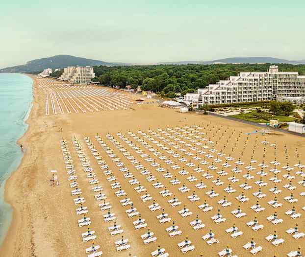 Ilmakuva suuresta hiekkarannasta, jolla on riveittäin aurinkotuoleja ja aurinkovarjoja sekä rakennuksia ja kasvillisuutta taustalla. (Tekoälyn luoma teksti)