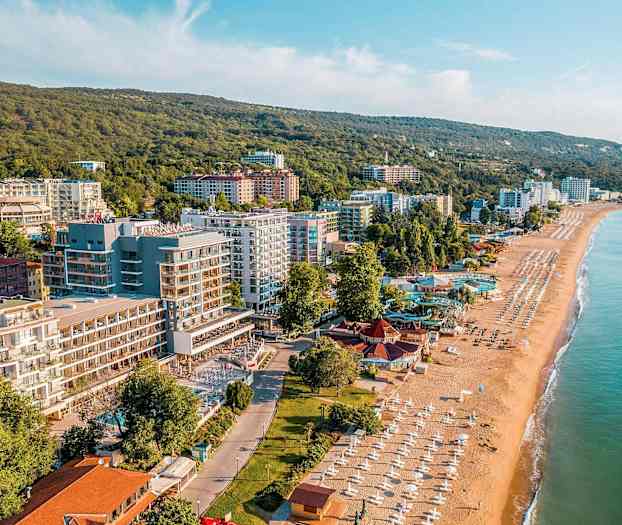 Kuva rannikkokaupungista, jossa hiekkaranta, aurinkovarjoja ja rakennuksia vehreiden mäkien vieressä. (Tekoälyn luoma teksti)