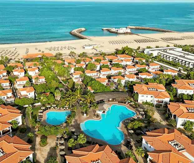 Ilmakuva lomakohteesta, jossa on valkoisia taloja oransseilla katoilla, uima-allas ja hiekkaranta meren rannalla. (Tekoälyn luoma teksti)