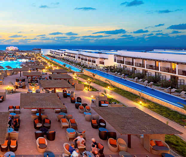 Hotellialue, jossa on uima-altaita, tyylikkäitä istuimia ja rakennuksia auringonlaskun aikaan. (Tekoälyn luoma teksti)