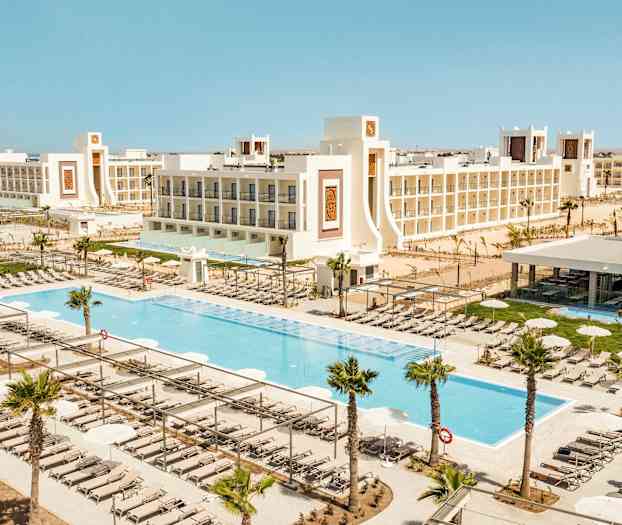 Näkymä lomakeskuksesta, jossa on uima-altaita, aurinkotuoleja ja valkoisia rakennuksia. (Tekoälyn luoma teksti)