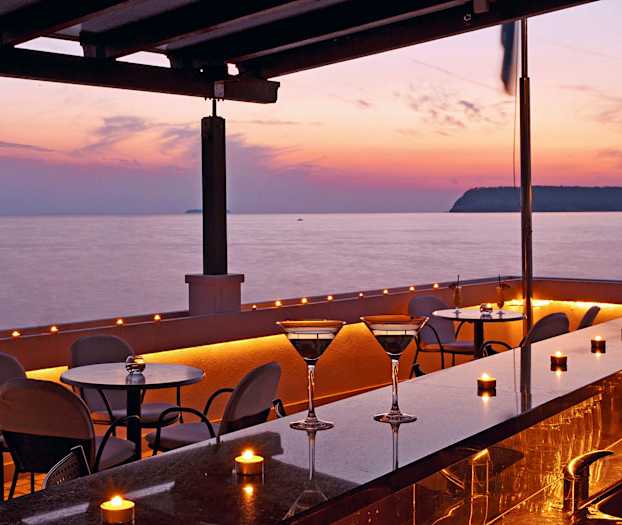 Ulkoterassi, jossa on pöytiä ja tuoleja meren rannalla auringonlaskun aikaan. (Tekoälyn luoma teksti)