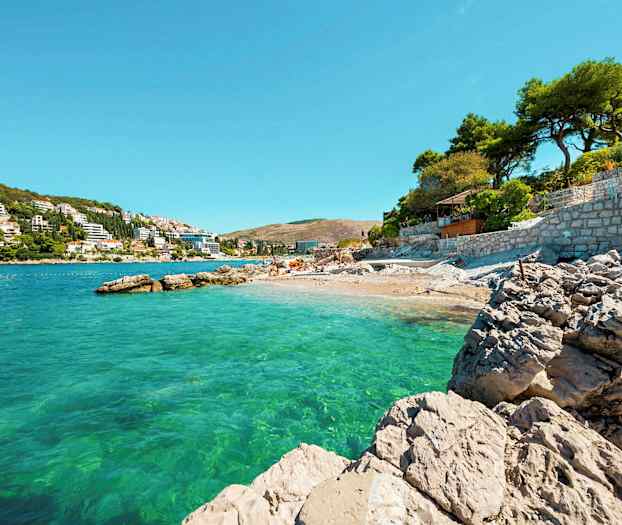 Kivinen ranta kirkkaan turkoosin meren äärellä, taustalla rakennuksia ja puita sekä kirkas sininen taivas. (Tekoälyn luoma teksti)