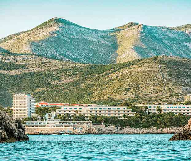 Rannikonäkymä, jossa merivesi on etualalla, rakennuksia keskellä ja vihreitä vuoria taustalla. (Tekoälyn luoma teksti)