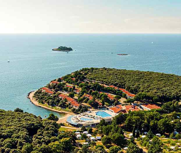 Ilmakuva niemestä, jossa on tiheää vehreyttä, punakattoisia taloja, uima-allasalue ja ympäröivä meri. (Tekoälyn luoma teksti)