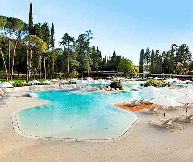 Ulkoallas, jossa vaaleansinistä vettä, ympäröitynä aurinkotuoleilla ja valkoisilla varjoilla vihreässä ympäristössä. (Tekoälyn luoma teksti)