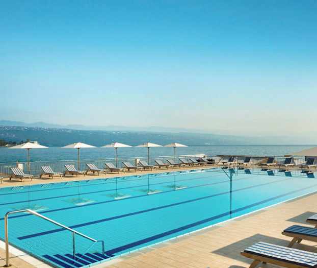 Ulkouima-allas, jonka reunoilla on aurinkotuoleja ja aurinkovarjoja. Taustalla näkyy meri ja vuoristo. (Tekoälyn luoma teksti)