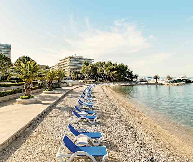 Kiviranta aurinkotuoleineen ja palmujen kera, hotellirakennusten ja puiden ympäröimänä. (Tekoälyn luoma teksti)