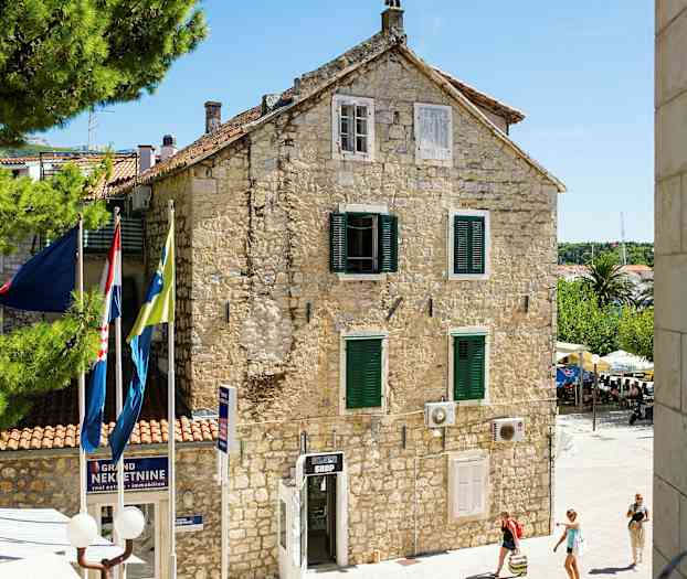 Kivitalo, jossa on vihreät ikkunaluukut ja lippuja edessä, ympärillä on puita ja ohikulkijoita. (Tekoälyn luoma teksti)