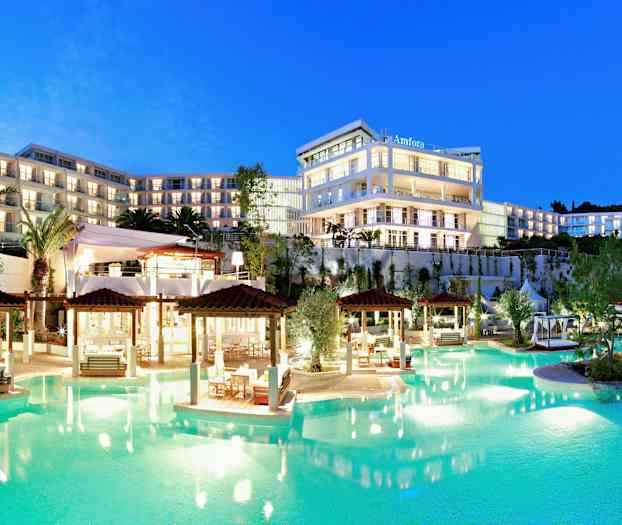 Valaistu monikerroksinen hotellikompleksi uima-altaan vieressä, ympäröitynä puutarhoilla ja palmuilla hämärtyvässä iltavalossa. (Tekoälyn luoma teksti)