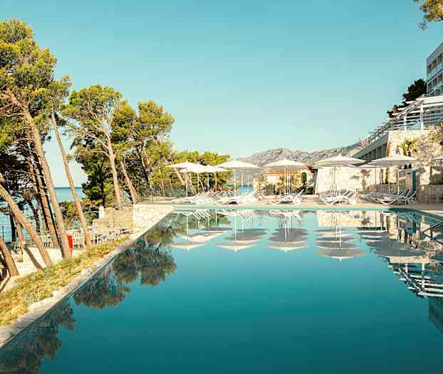 Näköala infinity-altaalle, jonka reunalla on aurinkotuoleja ja varjostimia, ympärillä puita ja rakennuksia. (Tekoälyn luoma teksti)