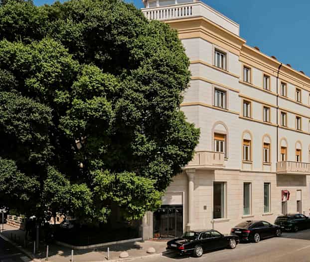 Usean kerroksen rakennus klassisella tyylillä, jossa on kaarevat ikkunat ja vaaleanvärinen julkisivu. Rakennuksen edessä on pysäköityjä autoja ja suuri vehreä puu. (Tekoälyn luoma teksti)