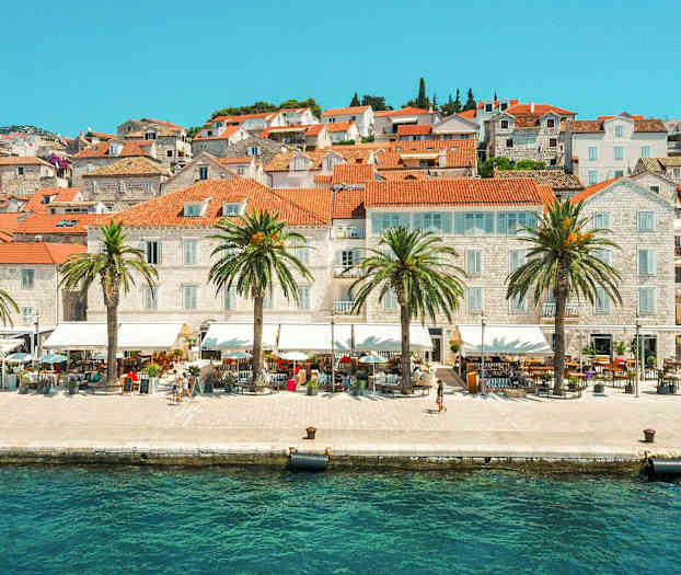 Kuva kaupungista veden äärellä, jossa on punatiilisiä kattoja ja palmuja rantakadulla. (Tekoälyn luoma teksti)