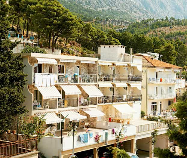 Valkoinen rakennus, jossa on terasseja, sijaitsee metsän reunalla ja vuorten lähellä. (Tekoälyn luoma teksti)