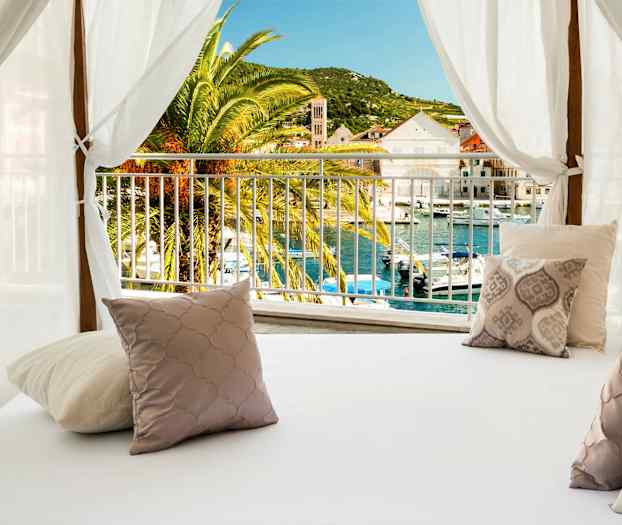 Vuode, jossa on koristetyynyjä valkoisen katoksen alla, näkymä palmuun ja satama. (Tekoälyn luoma teksti)