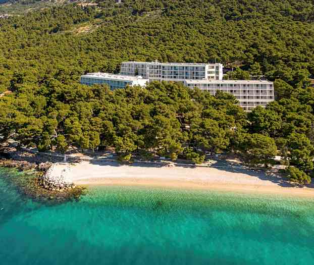 Hotellirakennukset vehreyden ympäröimänä lähellä rantaa, jossa on kirkasta turkoosia vettä. (Tekoälyn luoma teksti)