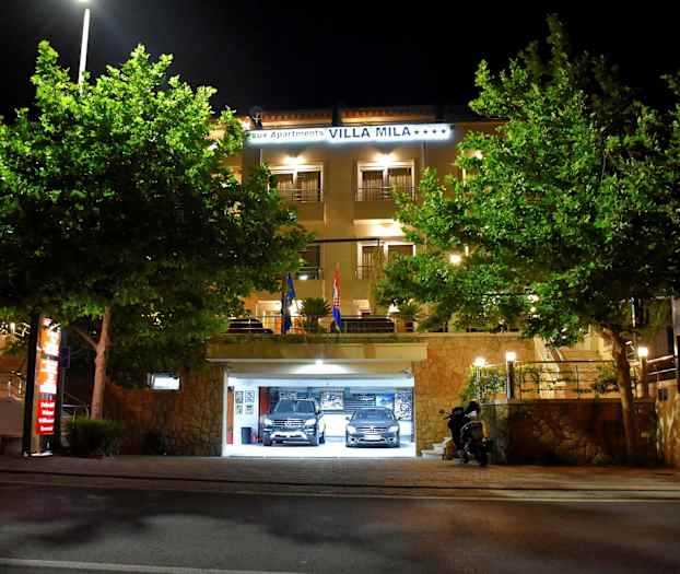 Hotellirakennus, jossa valaistu sisäänkäynti ja pysäköintihalli, ympäröitynä puilla, yöllä. (Tekoälyn luoma teksti)