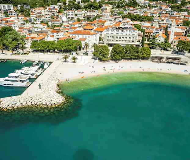 Ilmakuva rannasta, jossa on turkoosia vettä ja laitureita veneillä, ympärillä rakennuksia. (Tekoälyn luoma teksti)