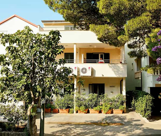 Kaksikerroksinen talo, jossa parvekkeet ja ilmastointilaitteet, ympäröitynä vehreydellä ja kukilla. (Tekoälyn luoma teksti)