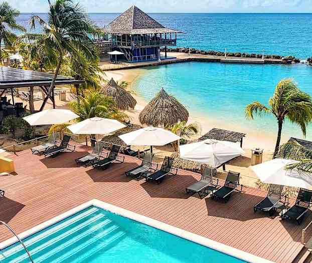 Näkymä rannalle, jossa on aurinkotuoleja, aurinkovarjoja, palmuja ja uima-allas. Taustalla näkyy meri. (Tekoälyn luoma teksti)