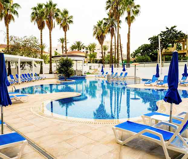 Kuva ulkouima-altaasta, jonka ympärillä on aurinkotuoleja, sinisiä aurinkovarjoja ja palmuja. (Tekoälyn luoma teksti)