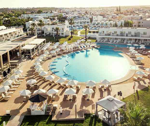 Suuri ulkouima-allas ympäröitynä aurinkotuoleilla ja aurinkovarjoilla, taustalla hotellirakennuksia. (Tekoälyn luoma teksti)