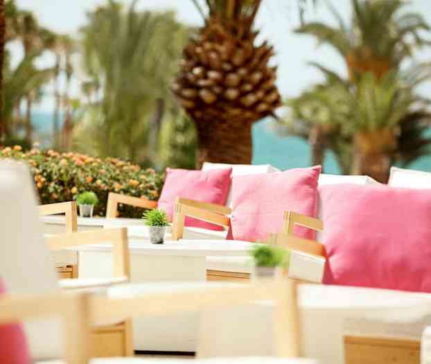 Ulkoterassi, jossa valkoiset istuinhuonekalut, koristeltuna vaaleanpunaisilla tyynyillä ja pienillä vihreillä kasveilla, ympärillä palmuja ja merinäköala. (Tekoälyn luoma teksti)