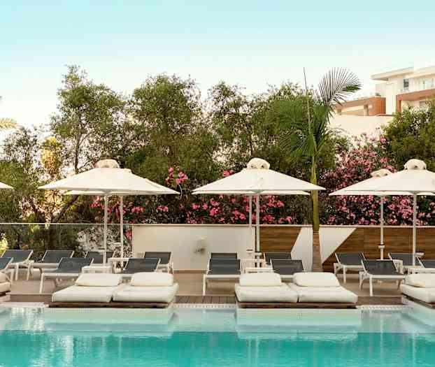 Kuva uima-altaasta, jossa on aurinkotuoleja ja aurinkovarjoja, ympäröitynä vehreällä kasvillisuudella ja vaaleansinistä taivasta. (Tekoälyn luoma teksti)