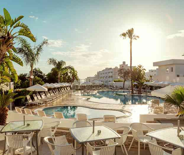 Kuvassa on hotellin ulkoallas, jota ympäröivät aurinkotuolit ja aurinkovarjot. Etualalla on pöytiä ja tuoleja aurinkoisessa säässä. (Tekoälyn luoma teksti)