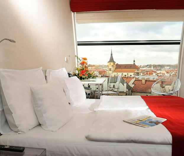 Hotellihuone, jossa on parivuode, koristeltu punaisella ja valkoisella vuodevaatteella. Panoraamanäkymä kaupunkiin, jossa on kirkon rakennuksia, näkyy ikkunan läpi. (Tekoälyn luoma teksti)