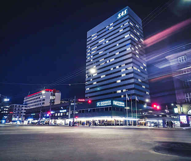 Korkea rakennus, jonka huipulla on SAS-logo, Radisson Collection -hotelli pohjakerroksessa, ympäröivät kaupunkirakennukset ja yöympäristö. (Tekoälyn luoma teksti)