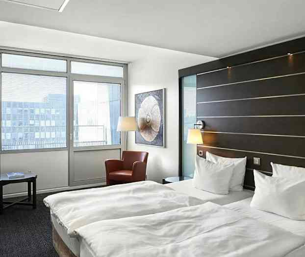 Hotellihuone, jossa on kaksi erillistä vuodetta, istumaryhmä kahdella nojatuolilla ja pöytä sekä suuret ikkunat, joista tulvii luonnonvaloa. (Tekoälyn luoma teksti)